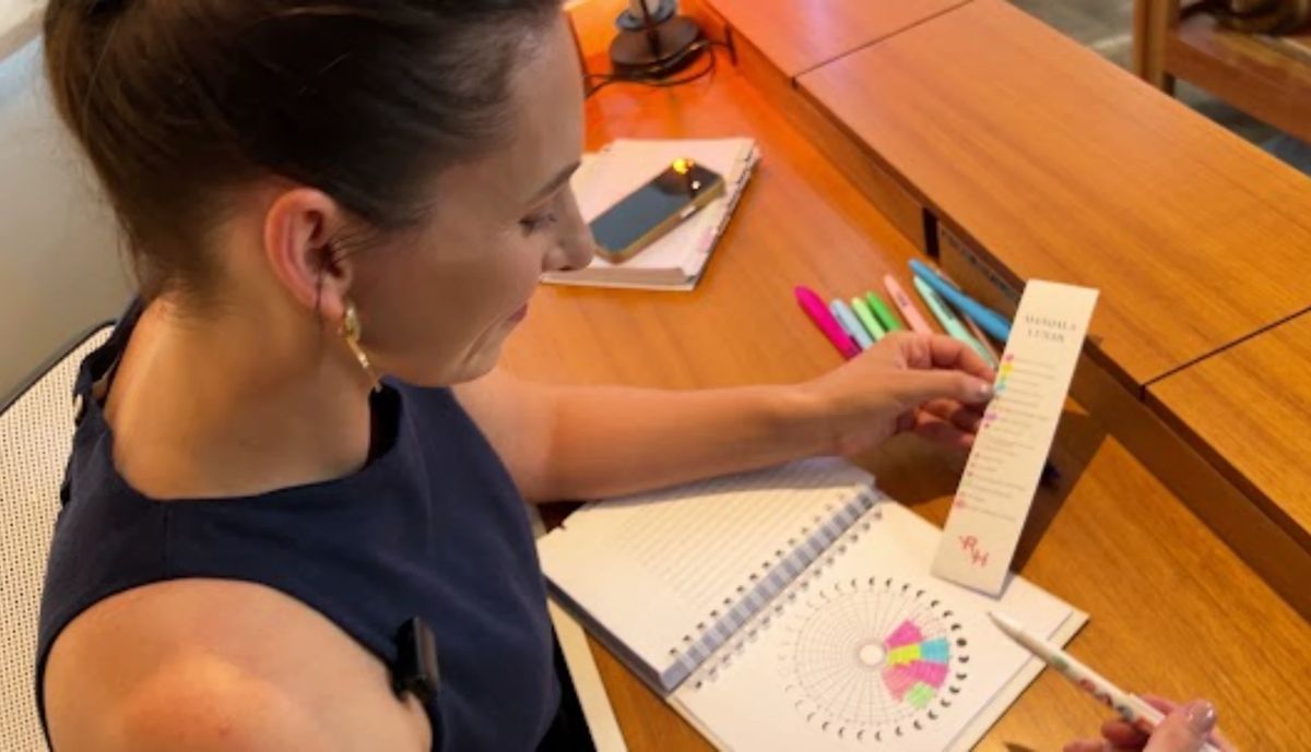 Renata Hermes à mesa, preenchendo uma mandala de luas no planner com canetas coloridas